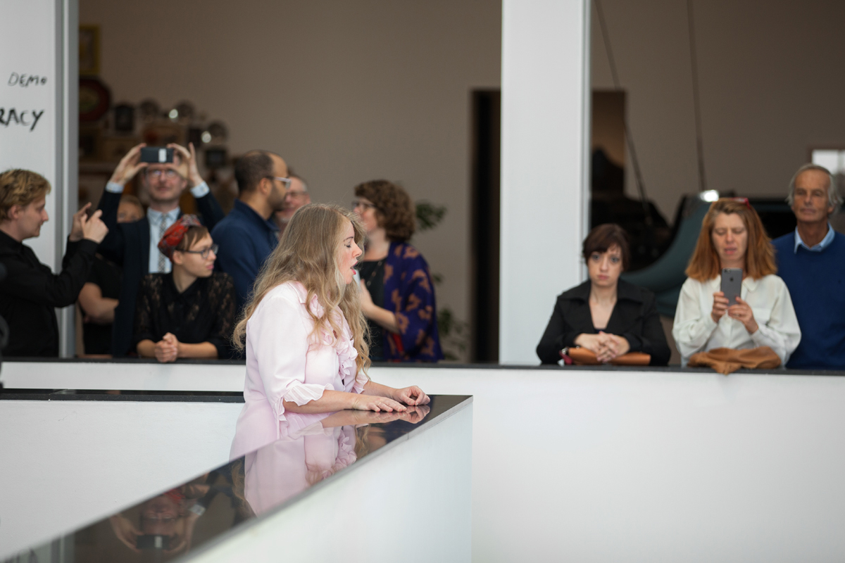 Color photograph of  the profile of a light-skinned woman singing and standing at the waist-high ledge inside a museum; in the background are numerous museum visitors watching her performance and taking photos on their phone