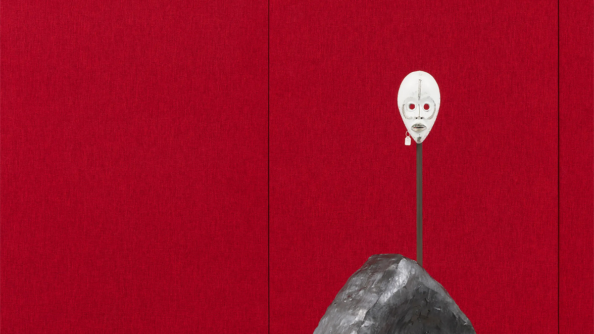 Color photograph of an artwork consisting of an African mask on a pole against a red wall