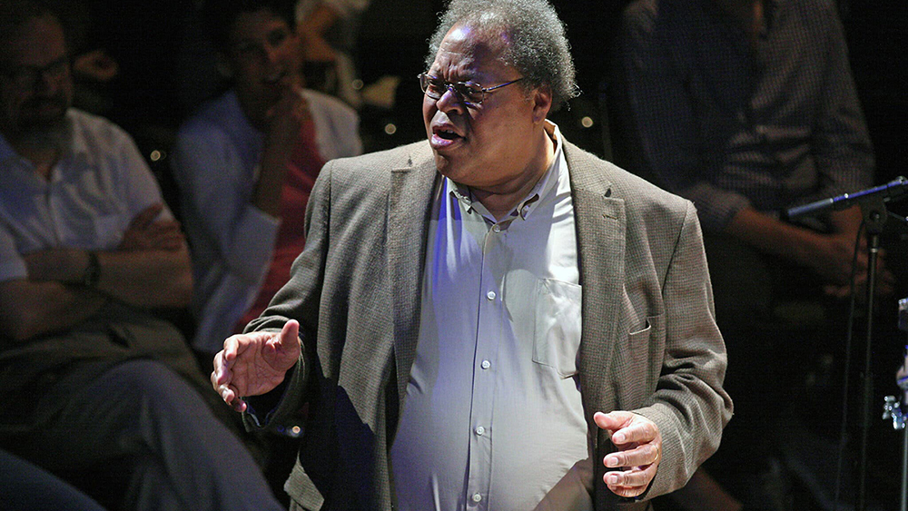 Color photograph of a dark skinned man with short white hair, wearing glasses, an open collar white shirt, and a gray blazer, standing in front of a seated audience and gesturing with both hands at waist level