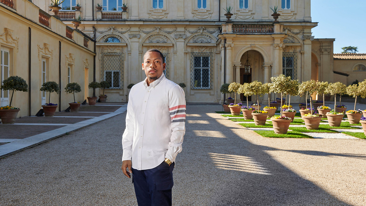 Color photograph of a dark skinned man wearing an untucked white button-up shirt and dark pants; he stands outdoors in front of a 17th-century Roman villa and looks directly at the camera