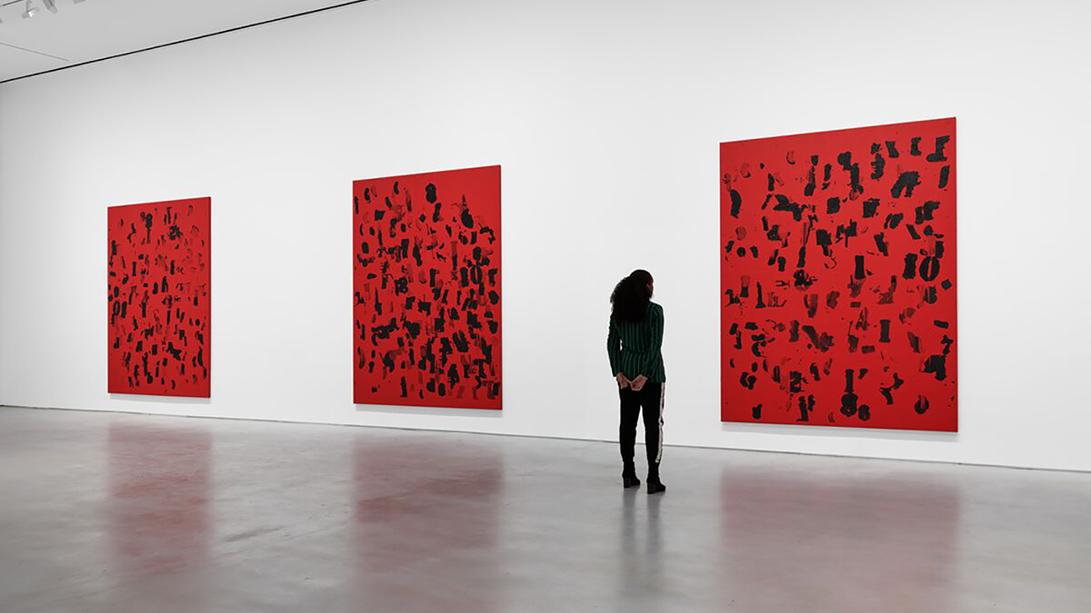 Color photograph of an installation of three square red and black paintings by Glenn Ligon in a white walled gallery with poured concrete floors; a dark skinned woman with black pants and top looks at one of them