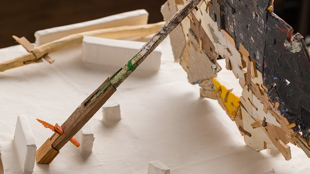 Color photograph of a close-up view of a tabletop sculpture by Helen O'Leary consisting of scraps of flat scraps of wood and linen standing upright but supported by a long straight dowel