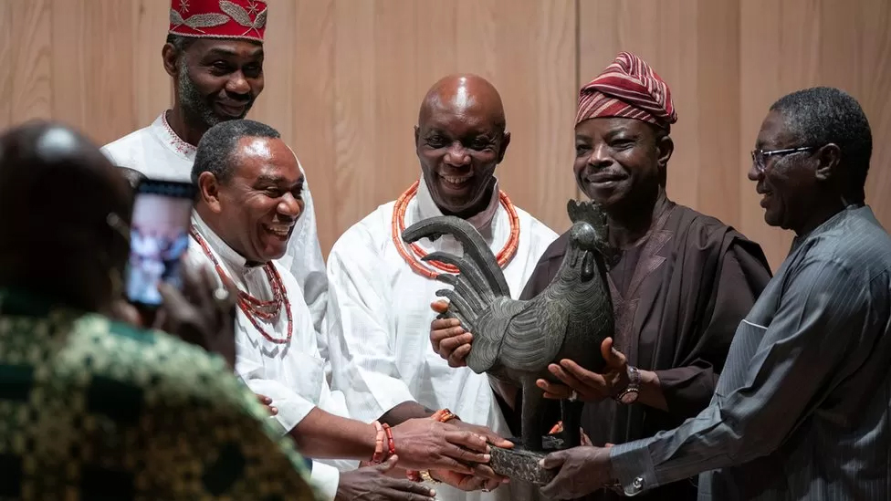Color photograph of six dark skinned men admiring a life sized bronze statute of a rooster