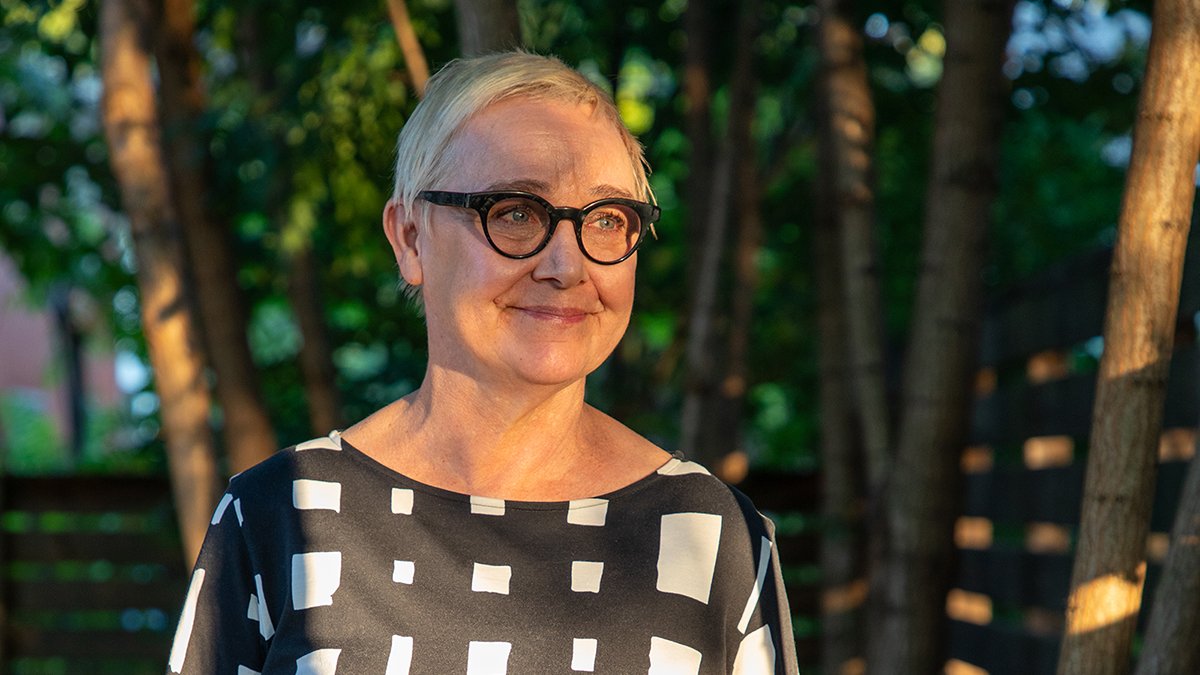 Color photograph of the head and torso of a woman with short gray hair and glasses, wearing a black and white dress and standing in front of trees, looking off to the side