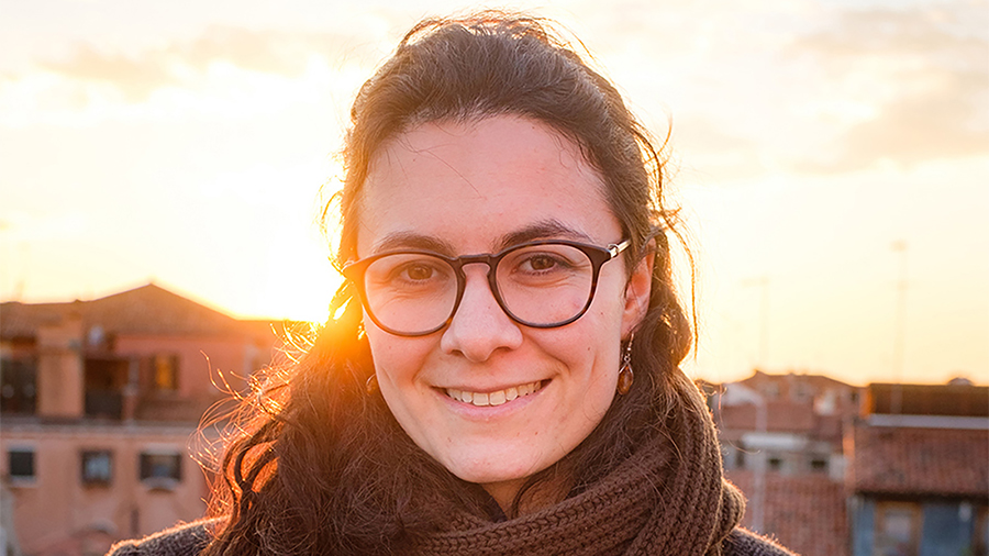 Color photograph of the head of a light skinned woman with brown hair and glasses, standing on an Italian rooftop or balcony, with the sun setting behind her