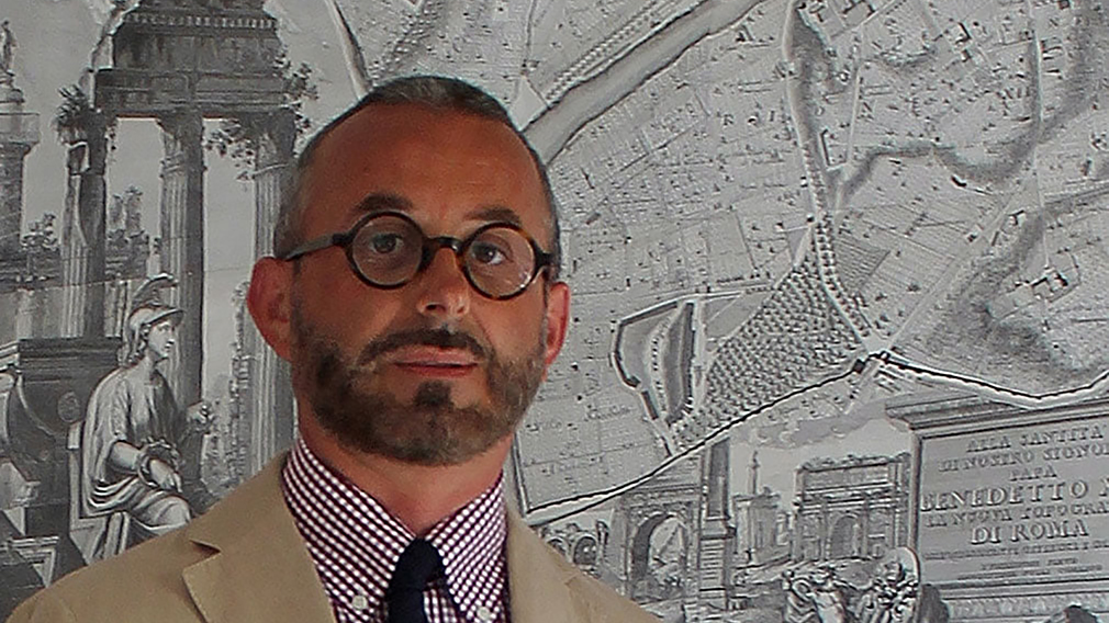Color photograph of the head and shoulders of an Italian man wearing round glasses and standing in front of a Noli map of Rome