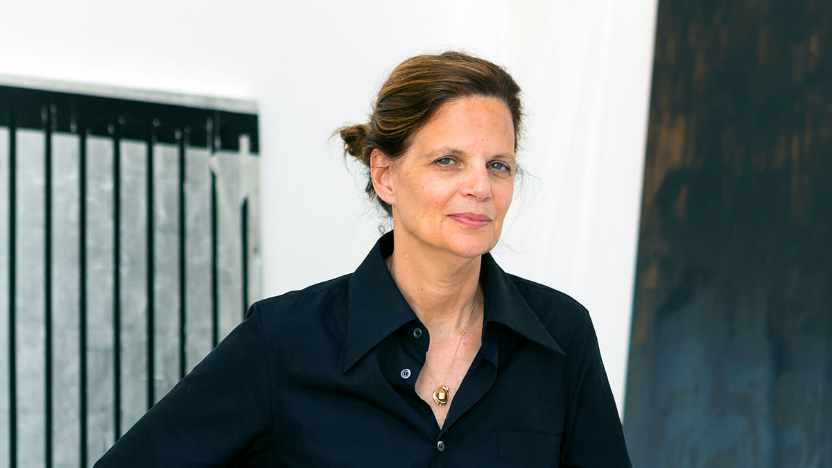 Color photograph of the head and torso of a light skinned woman wearing a dark open-collar shirt, looking at the camera with a slight but serious smile