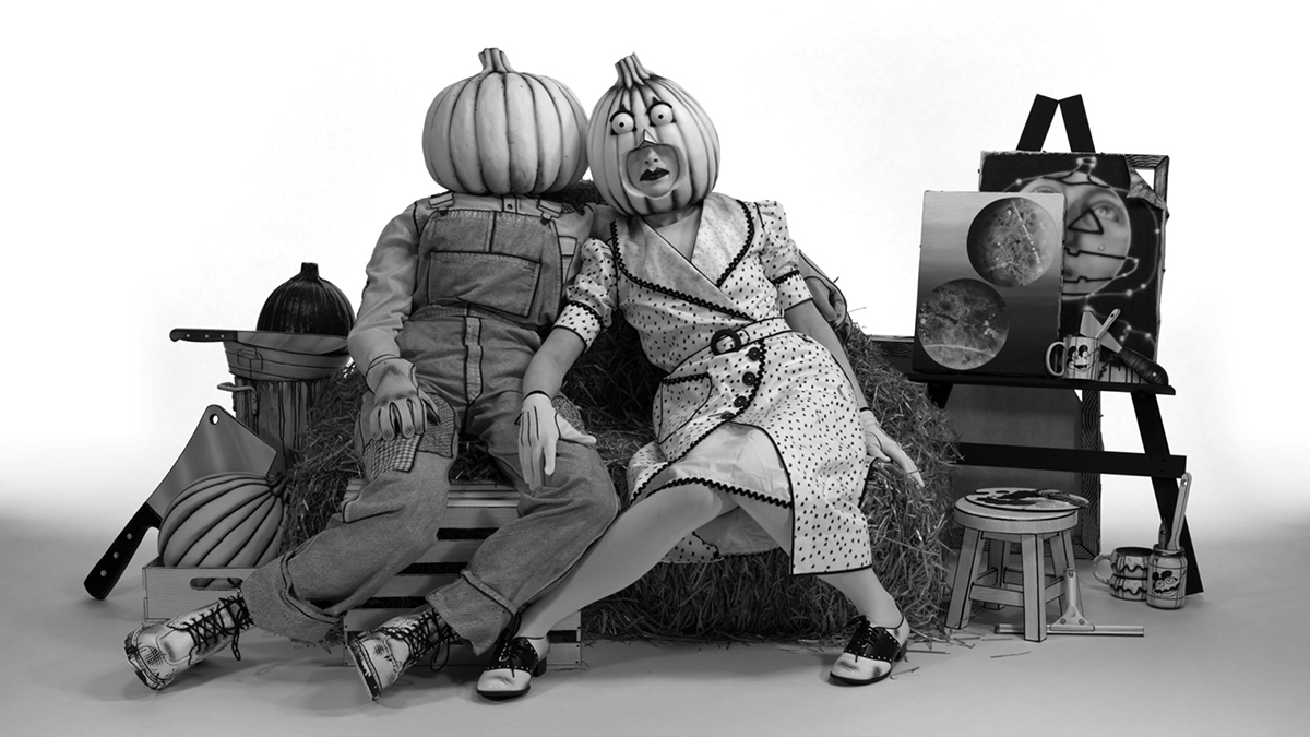 Still from a black and white art video showing two human figures sitting on a haystack bench, one in overalls and the other in a 1950s style dress. Both figures have pumpkin heads. To their left and right are scattered objects like a painter's easel and can of brushes, and a butcher's knife embedded in a third pumpkin