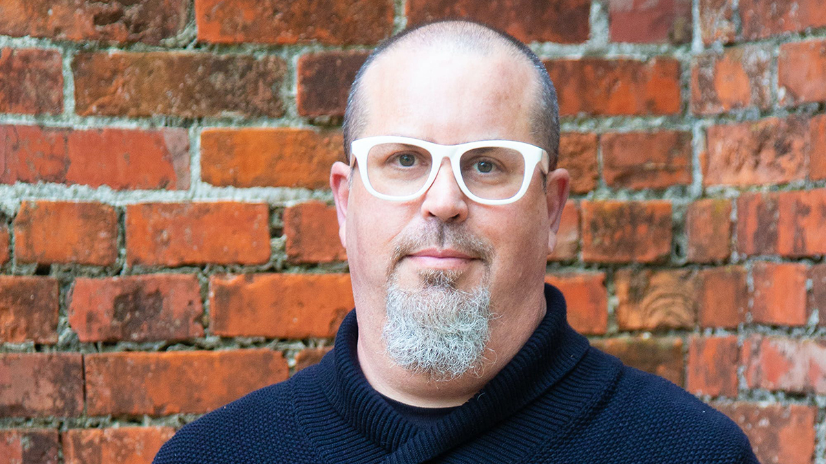 Color photograph of the head and shoulders of a light skinned man wearing glasses and a goatee; he stands in front of a red brick wall