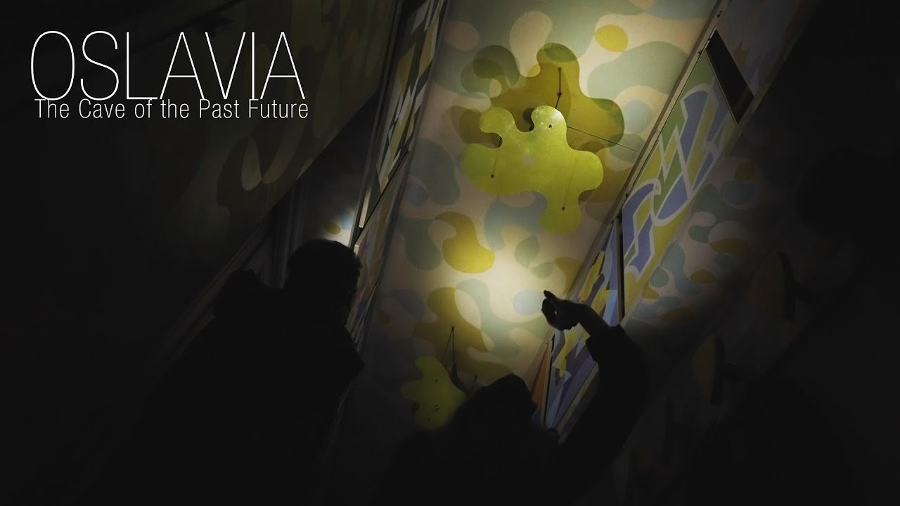 Still from the Ila Bêka and Louise Lemoine film showing hands holding flashlights in a darkened hallway, whose walls contain colorful abstractions. Spelled out in the upper left corner is the title: OSLAVIA - The Cave of the Past Future