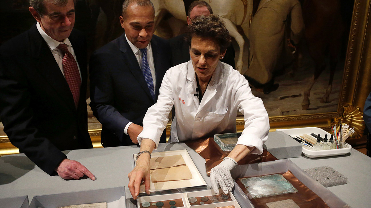 color photograph of a woman in a labcoat leaning over a table with objects as men stand behind her looking on