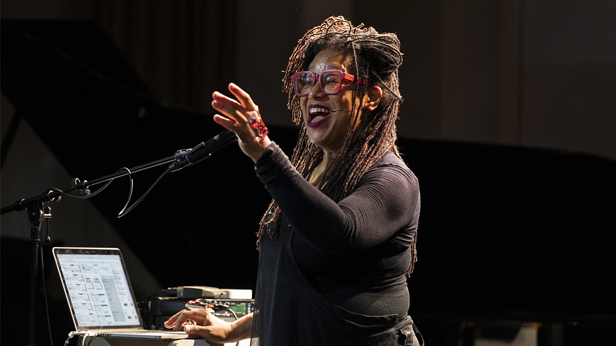 Color photograph of a black woman performing onstage; she raises her arm and opens her mouth as she uses a laptop with her other hand