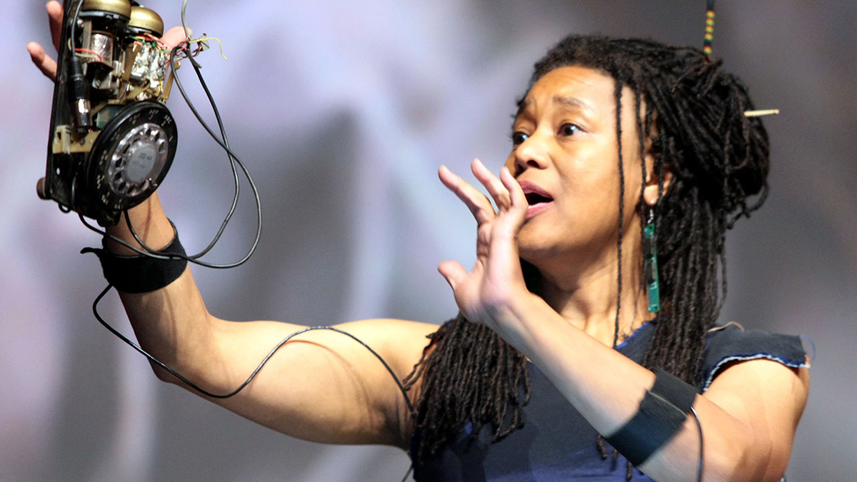 Color phorograph of a brown skinned woman performing onstage, she sings and holds up a hand built mechanical sound making device