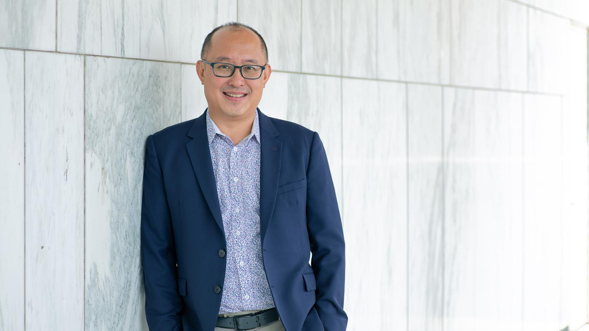Color photograph of an Asian man standing against a wall and wearing glasses and a blue blazer over a spotted blue and white button-up shirt; he smiles directly at the camera