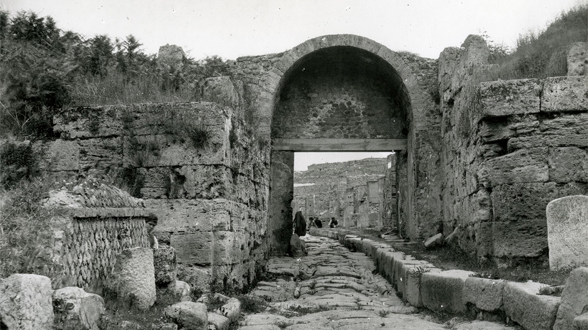 Black and white photograph from the early 20th century depicting a cobblestone road leading to an ancient Roman gate