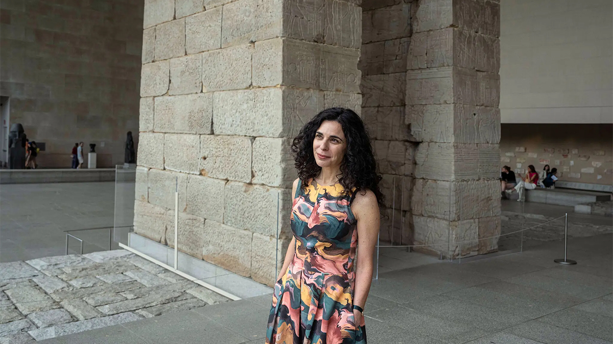 Color photograph of a woman with dark curly hair and a colorful dress standing near an Egyptian monument inside the Metropolitan Museum of Art