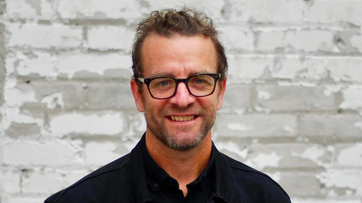 Color photo of the head and shoulders of a light skinned man wearing glasses and smiling at the camera in front of a white painted brick wall
