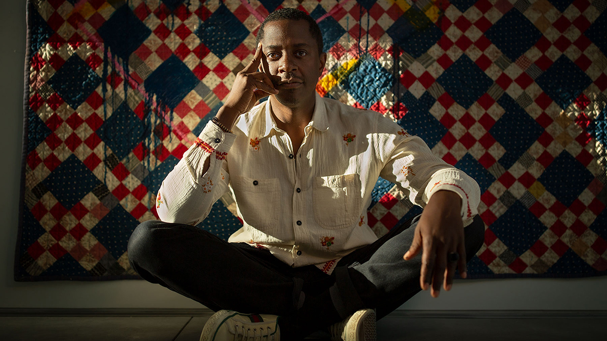 Color photograph of a dark skinned man sitting on the ground pretzel-style, with one elbow resting on his knee and the hand touching the chin; behind him is a colorful quilt of his own creation, with red, white, and blue squares dominating