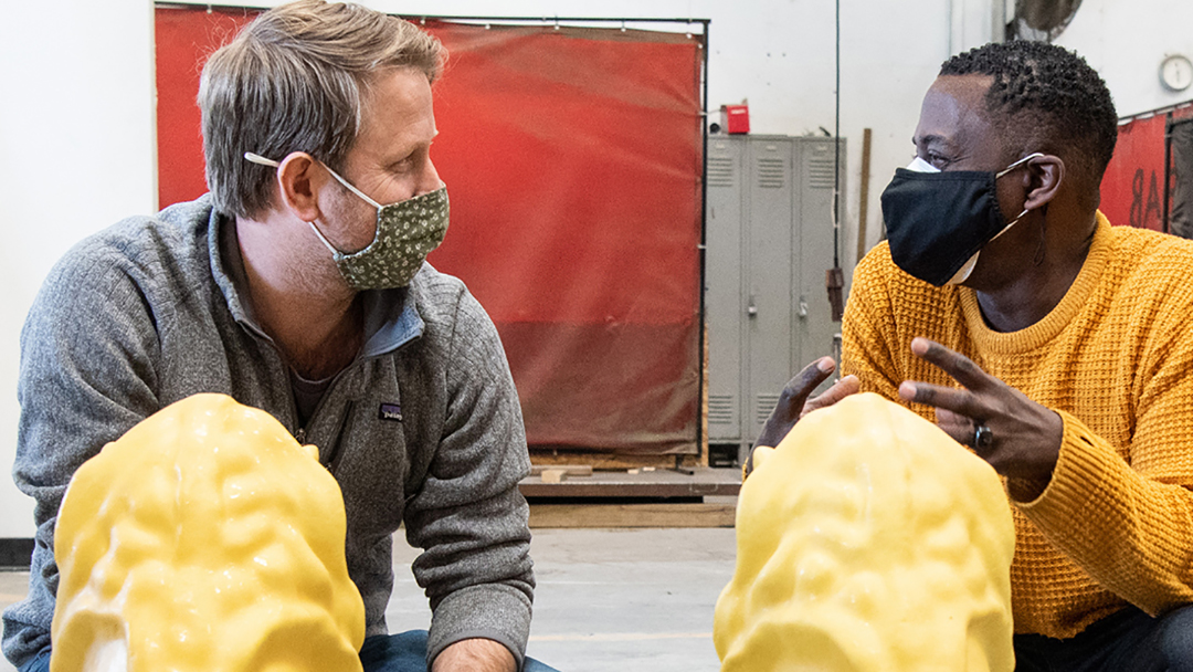 Color photograph of Sanford Biggers (on the right) crouching behind two yellow blobby sculptures beside another man (on the left); both men wear masks