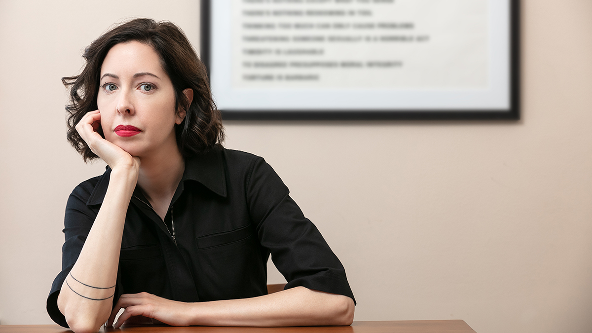 Color photograph of a light skinned, dark haired woman in a black top sitting at a table in an interior; she wears bright red lipstick and rests her chin on her right palm