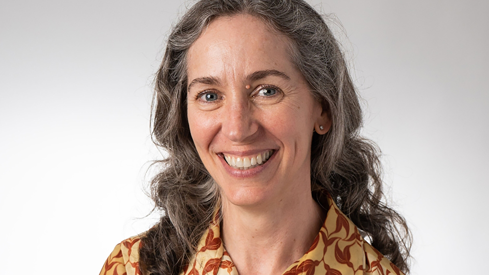 Color photograph of a light skinned woman with graying brown hair wearing a brown and yellow pattern blouse and smiling at the camera