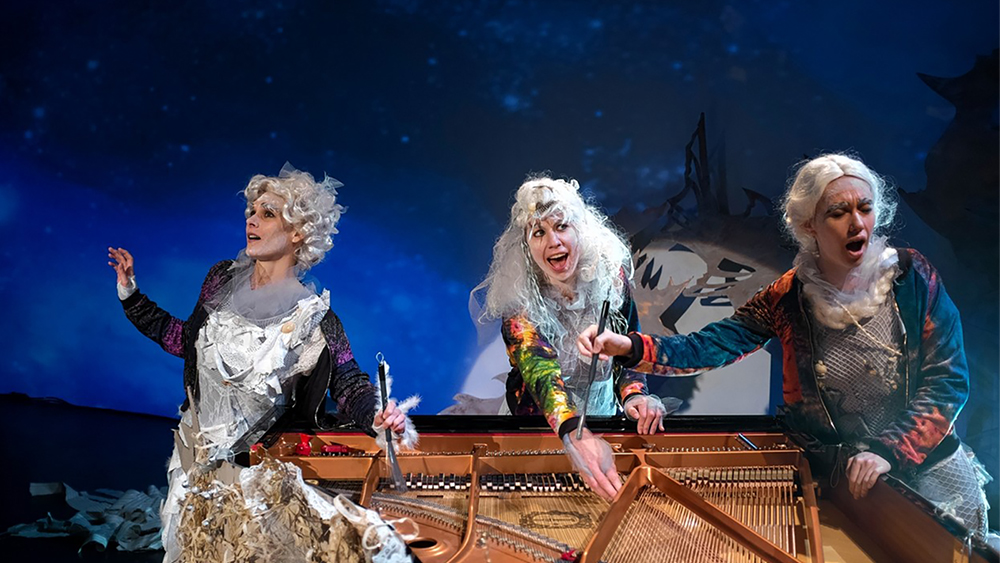 Color photograph of three women in siren costumes singing in front of a piano