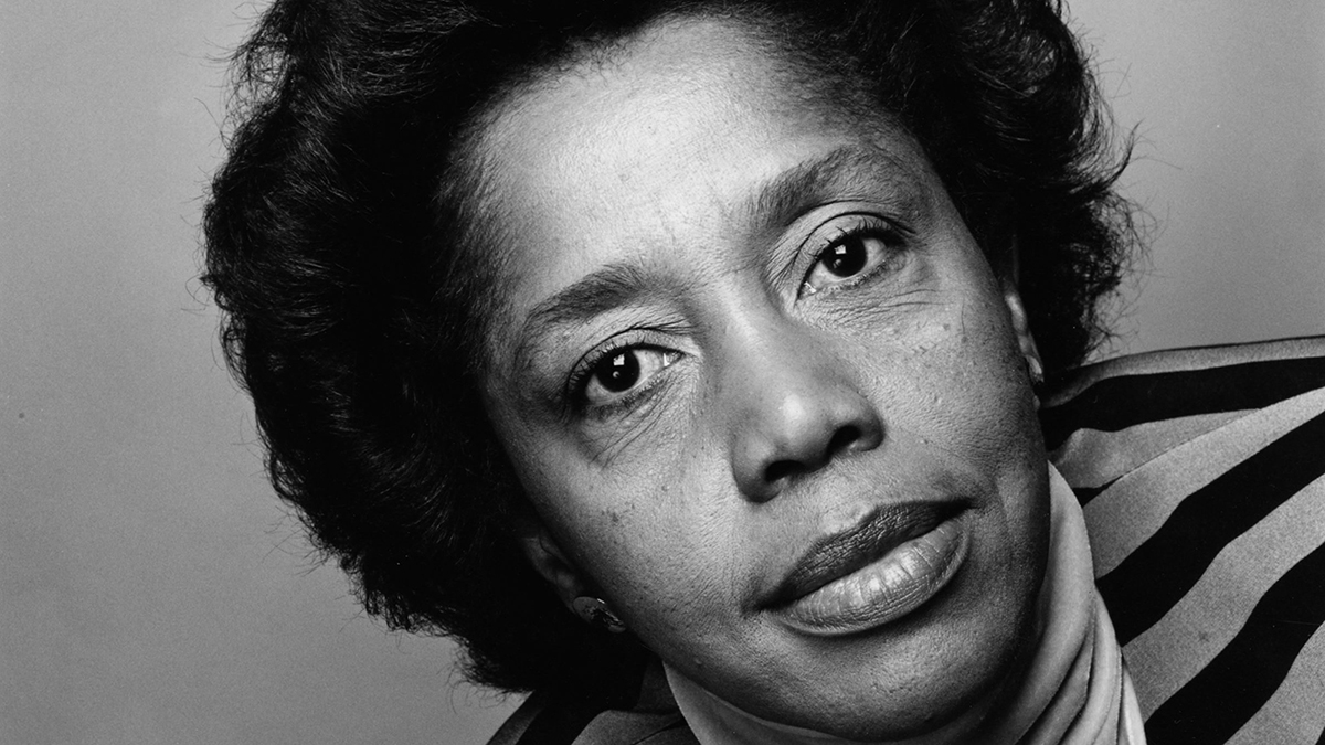 Black and white photograph of the head of dark skinned woman, titled to the left; she looks at the camera without smiling