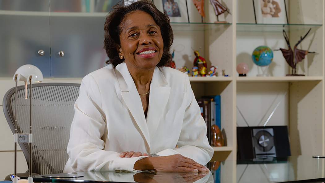 Color photograph of Tania León sitting indoors at a desk with a shelf of various items behind her