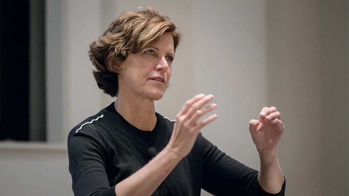 Color photo of the head and torso of a light skinned woman speaking and gesturing with her hands