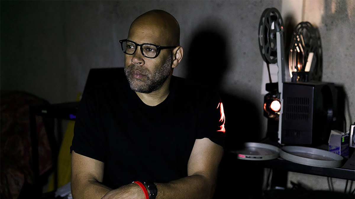 Color photograph of the head and torso of a dark skinned man sitting next to a film projector