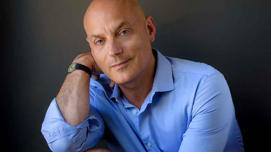 Color photo of the head and torso of a light skinned man wearing a blue collared button up shirt; he looks at the camera and smiles slightly