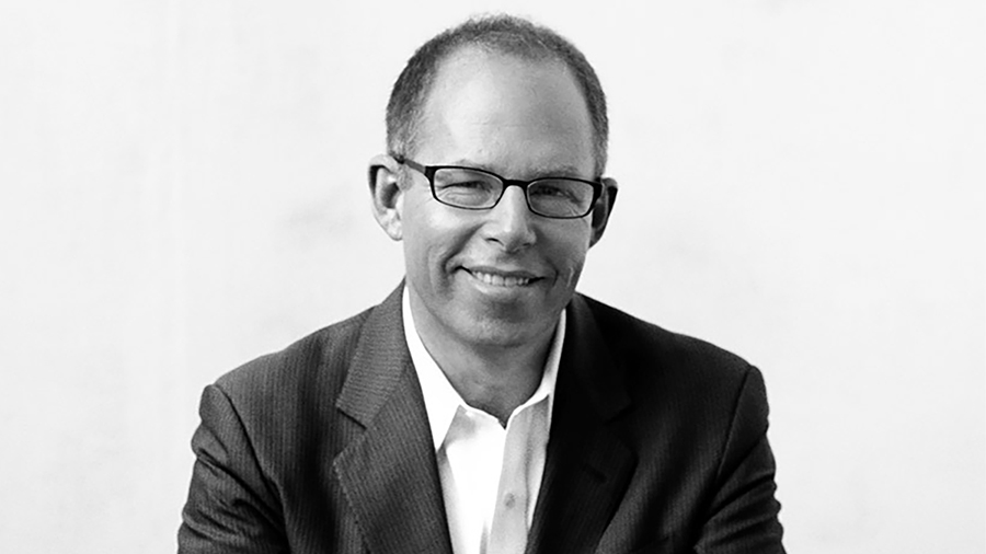 Black and white photo of the head and shoulders of a light skinned man wearing glasses and smiling at the camera