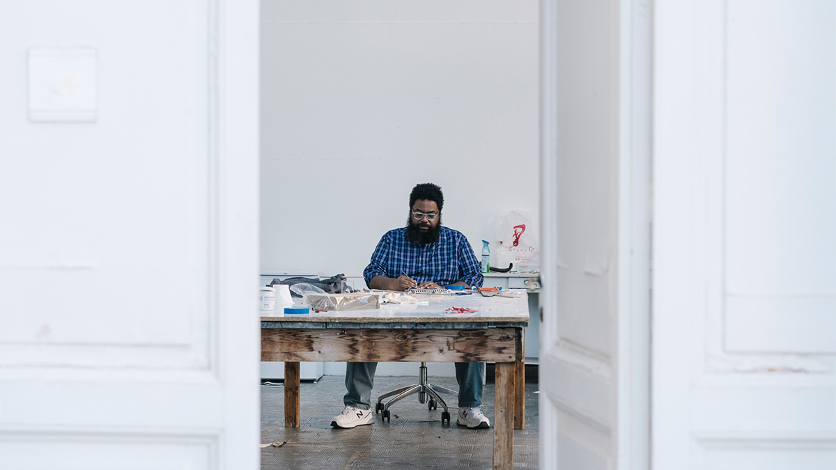 A dark skinned man sitting at a work table making art
