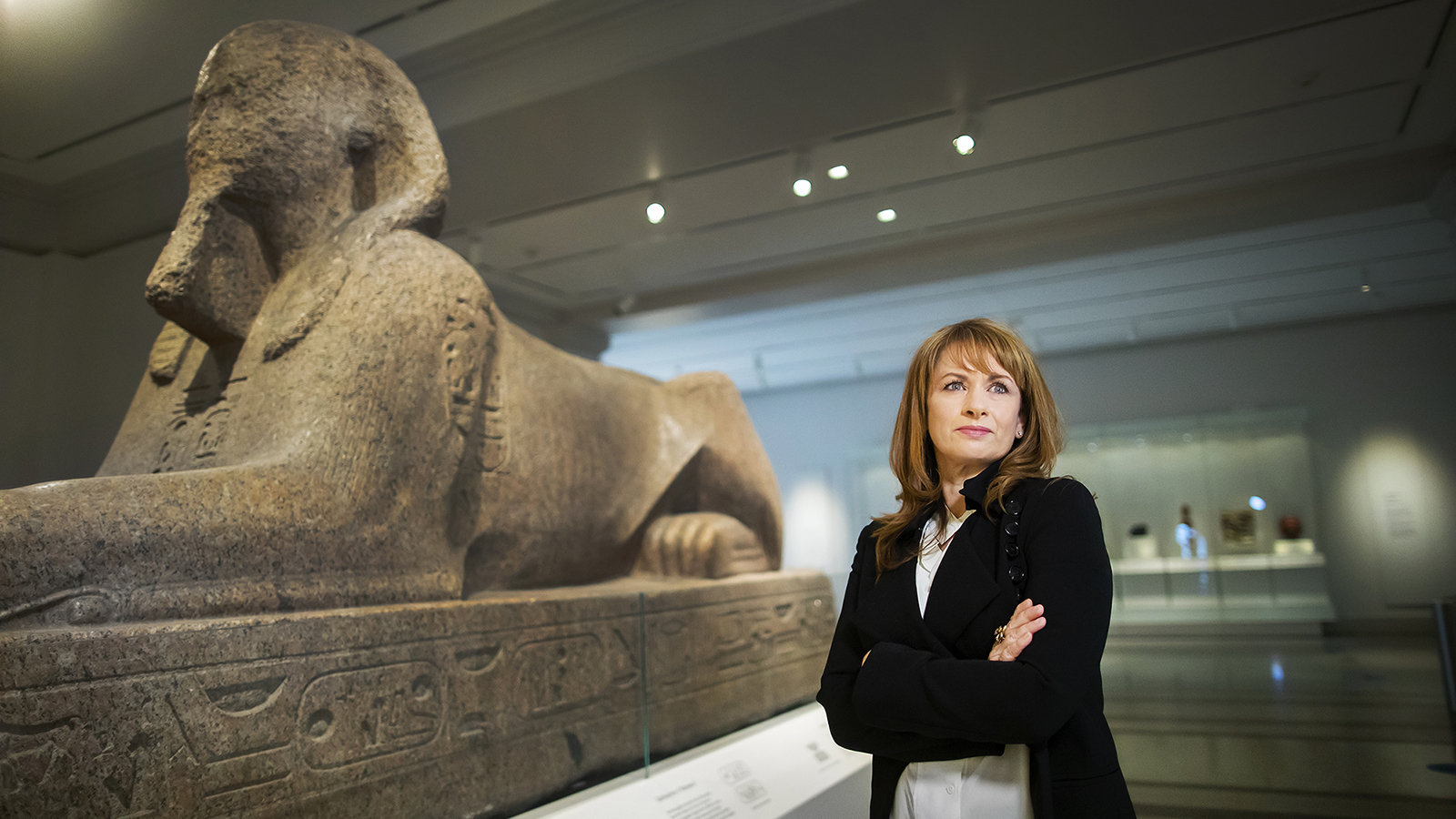 Color portrait of Lynne Meskell with her arms folded against her chest, inside a museum and standing next to an ancient sculpture of a sphinx