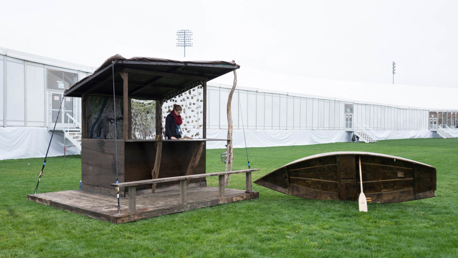 Color photograph of a person standing in a wooden stall on the grass, next to a wooden rowboat on its side