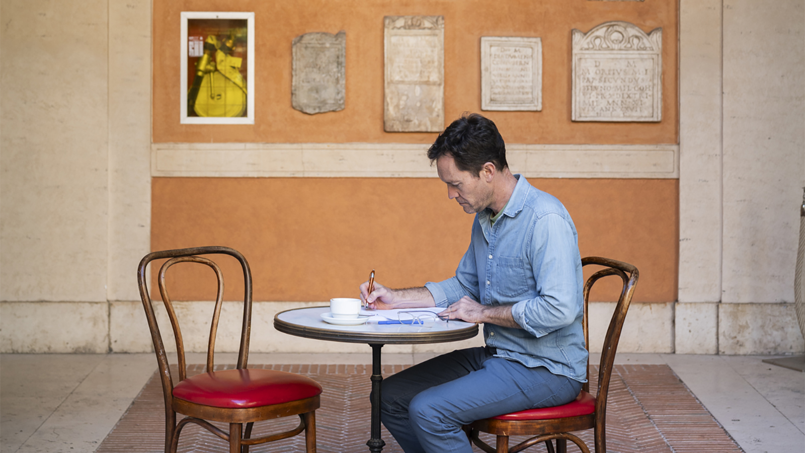Color photo of a light skinned man seated at a cafe table and writing in a notebook