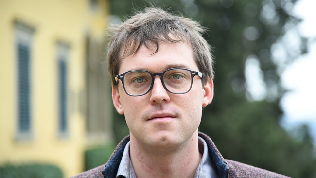 Color photograph of the head of a light skinned man wearing glasses, standing outside a building and looking directly at the camera