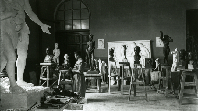 Black and white photograph of an artist's studio depicting a sculptor working among many examples of his figurative work