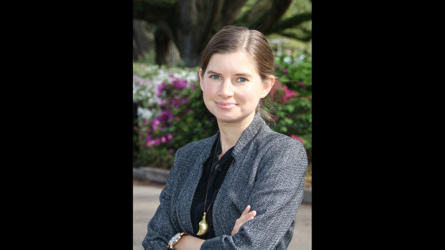 Color photograph of the head and torso of a light skinned woman wearing a gray blazer; she stands with her arms crossed and smiles slightly at the camera