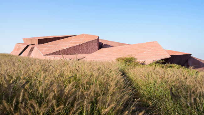 color photograph of a jagged modern brown building, with a wheat field in front of it