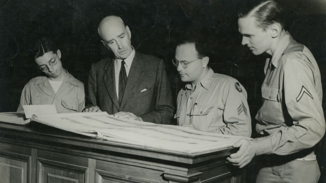 Black and white photo of three white men and one white woman looking at a large book