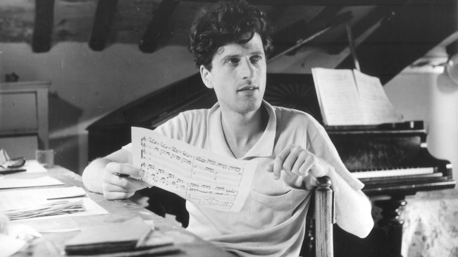 Black and white photograph from 1952 of a light skinned man in a short sleeve shirt seated at a table; he holds a page of sheet music; a piano can be seen behind him