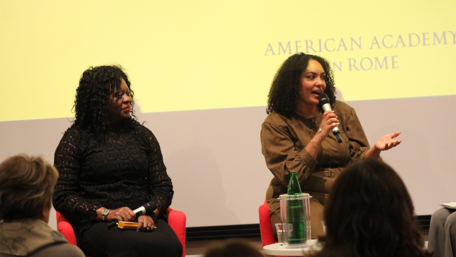 Two Black women seated in chairs in a lecture room; one of them is speaking into a microphone