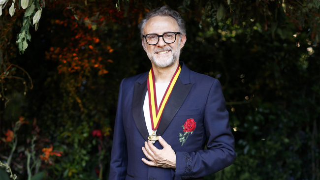 Color photograph of a light skinned man wearing glasses, a white shirt, and a blue suit; in his hand he holds up the medal that is draped around his neck