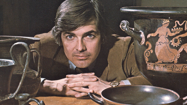 Color photograph ca. 1960s or 1970s of the head, shoulders, and folded hands of a light skinned man in a brown sportscoat posing above a tabletop of Greek vases and pottery