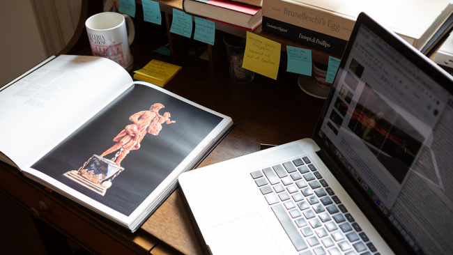 Color photo of a desk with an open laptop, an open book, and sticky notes