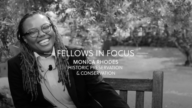 Black and white screeshot of a video depicting a dark skinned woman sitting in a garden, wearing a dark blazer, and smiling at the camera