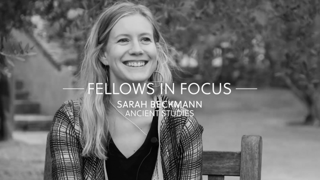 Black and white video still of the head and torso of a smiling light skinned woman with blond hair sitting in a chair in a garden; superimposed onto her is a text reading "Fellows in Focus: Sarah Beckmann, Ancient Studies"