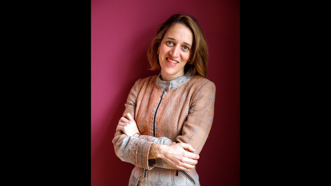 Color photograph of a light skinned woman standing with her arms crossed and smiling at the camera in front of a fuscia wall