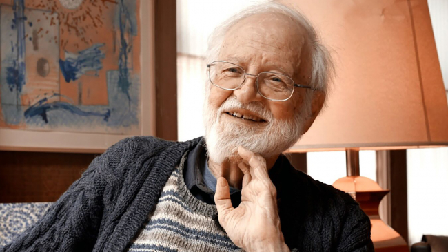 Color photograph of the head and shoulders of an elderly light skinned man wearing a sweater and glasses and seated in a living room; one hand touches his white beard as he looks off camera